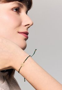 Zielona bransoletka z kordelki z złotym kwiatowym charms oraz akcentami w postaci koralików, noszona na jasno skórze, podkreślająca jej delikatny design.