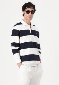 Randig långärmad polo i marinblått och vitt med krage och logotyp på bröstet, tillsammans med ljusa byxor. Modellen bär solglasögon.