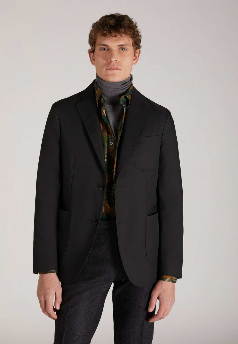 Blazer nero con rever a punta e una tasca sul petto, indossato sopra una camicia fantasia e un dolcevita grigio. Tessuto testurizzato, vestibilità sartoriale.