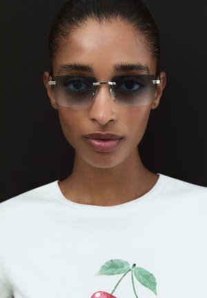 Occhiali da sole con montatura grigia trasparente, forma rettangolare e dettagli metallici. Indossati con una camicia bianca con stampa di ciliege.