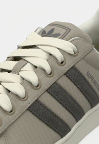 Baskets beiges avec une tige en toile texturée, des lacets crème, des bandes en daim gris et un logo embossé sur la languette, avec des œillets ronds.