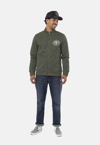 Veste en polaire zippée vert olive avec un patch sur le côté gauche de la poitrine, associée à un jean bleu et des chaussures à enfiler à carreaux noir et blanc.