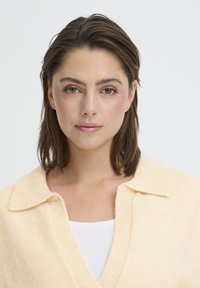 Pull tricoté couleur crème avec un col en V et un large col. Texture douce, avec des détails côtelés, porté par-dessus un haut blanc.