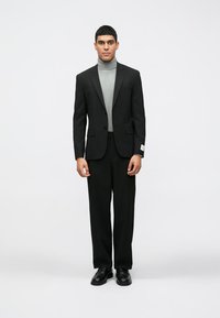 Melnas vīriešu uzvalks ar diviem pogām, bez raksta; kombinēts ar pieguļošu pelēku turtleneck. Melnas ādas kurpes pabeidz tērpu.