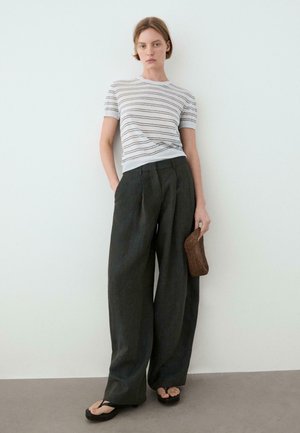 Frau in weißem, kurzärmeligem, gestreiftem Oberteil, lockeren dunkelweiten Hosen, schwarzen Sandalen, hält eine kleine braune geflochtene Clutch, steht vor einer weißen Wand.