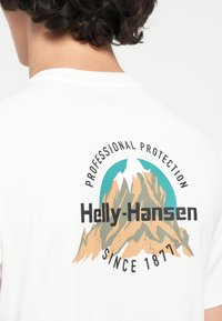 Pogled sa leđa osobe koja nosi bijelu košulju s Helly-Hansen logom, grafikom planine i natpisom "Profesionalna zaštita od 1877."
