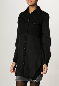 Camicia nera con bottone, maniche lunghe, due tasche sul petto, texture liscia e orlo curvo, indossata sopra una gonna di jeans.