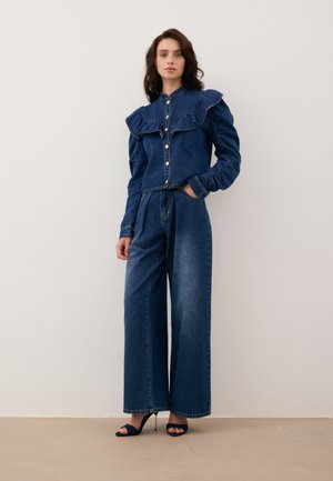 Denimoutfit met een knoopjestop met een gerimpelde kraag en wijde broekspijpen. Diepblauwe kleur met een gladde textuur. Hakken.