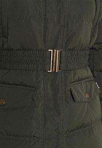 Veste matelassée vert foncé avec ceinture élastique munie d'une boucle métallique et deux poches à rabat avec boutons-pression.