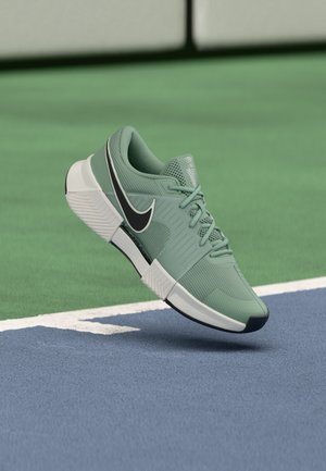 Zeleni športni čevelj Nike z belo in črno podplatom, ki lebdi nad modro-zelenim tenis igriščem blizu bele črte.