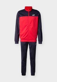 Κόκκινη και ναυτική φόρμα από λείο ύφασμα· διαθέτει ζακέτα με φερμουάρ και στενό γιακά, καθώς και παντελόνια με ελαστική μέση και διακοσμητικά λογότυπα.