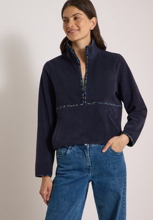 Kvinde iført en marineblå fleece-trøje med kvart lynlås og camouflagekant, blå jeans, stående med den ene hånd i lommen og med et let smil.