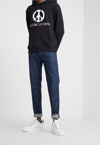 Svart hoodie med ett stort vitt fredssymbol och flerfärgad "LOVE MOSCHINO" text, kombinerat med blå denimjeans och vita sneakers.