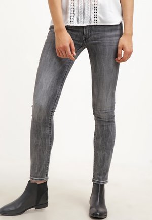 Person iført falmede grå skinny jeans, sorte ankelstøvler og en hvid top med blonde detaljer, stående mod en ensfarvet baggrund.
