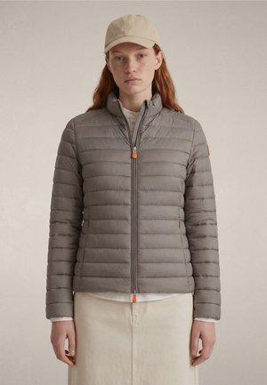 Young woman with red hair wears light gray puffer jacket, beige cap, white shirt, and cream skirt, facing forward against plain background.