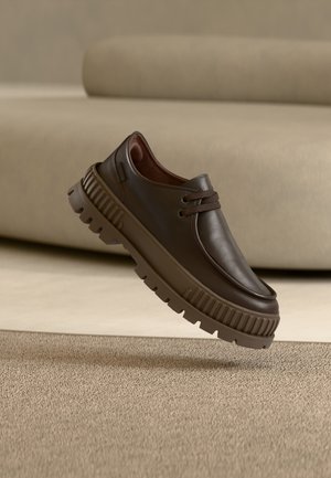 Chaussure décontractée en cuir marron foncé avec semelle épaisse et texturée et lacets, suspendue au-dessus d'un tapis beige dans une pièce aux tons neutres.
