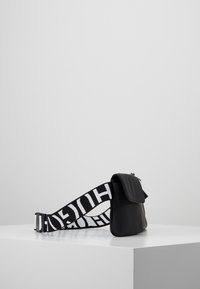 Borsa nera con superficie testurizzata, caratterizzata da un classico lembo piegato e una larga tracolla contrassegnata da lettere bianche in grassetto. Design compatto e rettangolare.
