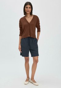 Cardigan marron à col en V avec des boutons, associé à un short bleu foncé à cordon de serrage. Le modèle porte des chaussures slip-on beiges. Texture de tissu lisse dans l'ensemble.