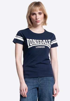 Mujer rubia vistiendo una camiseta azul marino de Lonsdale London con rayas blancas en las mangas y jeans azules, posando frente a un fondo blanco.