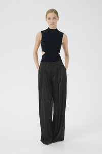 Schwarzes ärmelloses Oberteil mit Ausschnitten, kombiniert mit hochgeschnittenen schwarzen plissierten Hosen, die eine moderne, minimalistische Silhouette schaffen.