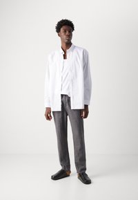 White and gray striped shirt, white tank top underneath, gray linen pants, and black clogs. Model stands upright on a white background.