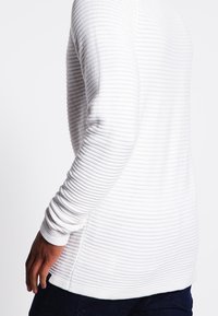 Maglione bianco a maniche lunghe con una texture a coste e sottili righe verticali, dotato di maniche aderenti e scollatura rotonda.