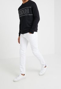 Schwarzer Pullover mit Logo auf der Brust, kombiniert mit weißen Jeans in Slim-Fit und weißen Turnschuhen. Glatte Texturen, lässiges Design.