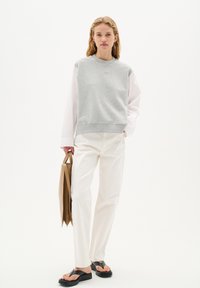Felpa grigia con polsini a costine sopra una maglietta a maniche lunghe bianca, abbinata a pantaloni color crema e sandali neri, mentre tiene una borsa marrone.