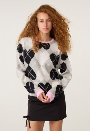Jonge vrouw met krullend rood haar draagt een grijze argyle trui met zwarte harten en roze manchetten, gecombineerd met een zwarte rok met een kleine strik aan de zijkant.