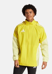 Veste imperméable jaune avec des accents blancs, zip sur le devant, capuche réglable et poignets élastiques. Affiche le logo Adidas sur la poitrine.