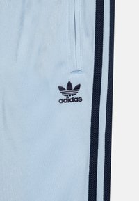 Šviesiai mėlyni sportiniai džinsai su tamsiai mėlynu šoniniu raiščiu ir juodu Adidas logotipu siuvinėjimu kairiajame šlaunyje. Lygus paviršius.