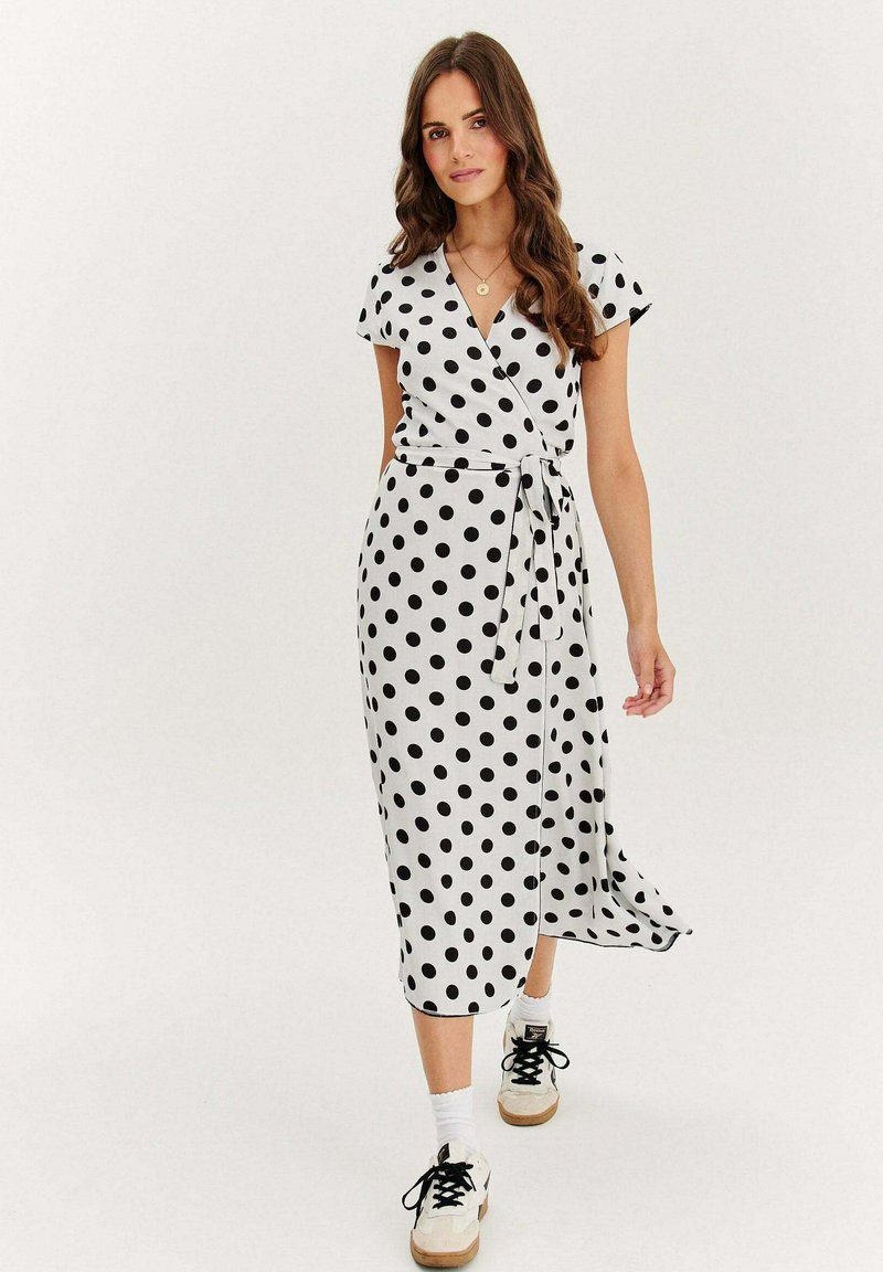 Černobílé puntíkované šaty s krátkým rukávem, zavazovací pas, délka ke kolenům, vyrobené z hladké látky, doplněné bílými teniskami.