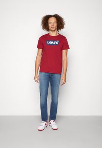 Rød bomull t-skjorte med en blå Levi's-logo, kombinert med lyseblå jeans og hvite sneakers med røde detaljer. Enkelt design og passform.