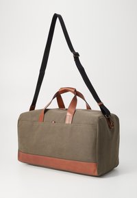 Olive green canvas duffel bag with brown leather accents, dual top handles, and a detachable black shoulder strap. Features a zip closure.