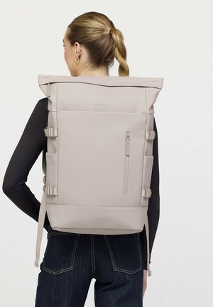 Frau mit blondem geflochtenem Pferdeschwanz, die einen beigen Rolltop-Rucksack und ein schwarzes Langarmshirt mit dunkler Jeans trägt, steht mit dem Rücken zur Kamera.