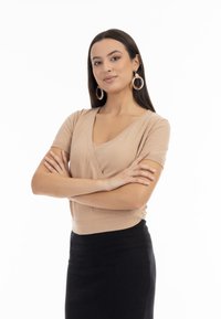 Krémová krátká halenka s výstřihem do V a vypasovaným pasem, v kombinaci s černou sukní. Modelka nosí velké kruhové náušnice. Hladká textura látky.