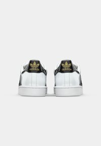 Tenisky Adidas s lesklým bielym koženým povrchom, čiernymi akcentmi a zlatým logom na päte. Obsahujú bielu gumovú podrážku a textúrované pruhy.