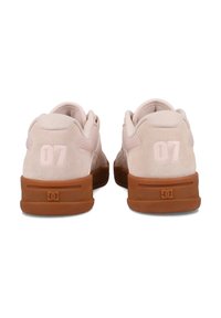 Een paar lichtroze suède sneakers met gumzolen, nummer "07" op de hakken, en het DC-logo op de achterkant van de bruine rubberen zolen.