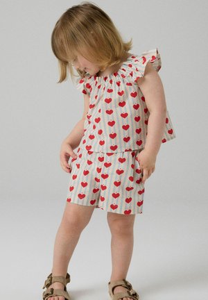 Petite fille portant un haut assorti à motifs de cœurs avec manches à volants et un short, regardant vers le bas avec une main dans la poche.