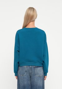 Sudadera de color verde azulado con cuello redondo, hombros caídos y dobladillo acanalado. Combinada con jeans de denim de ajuste holgado de color azul claro.