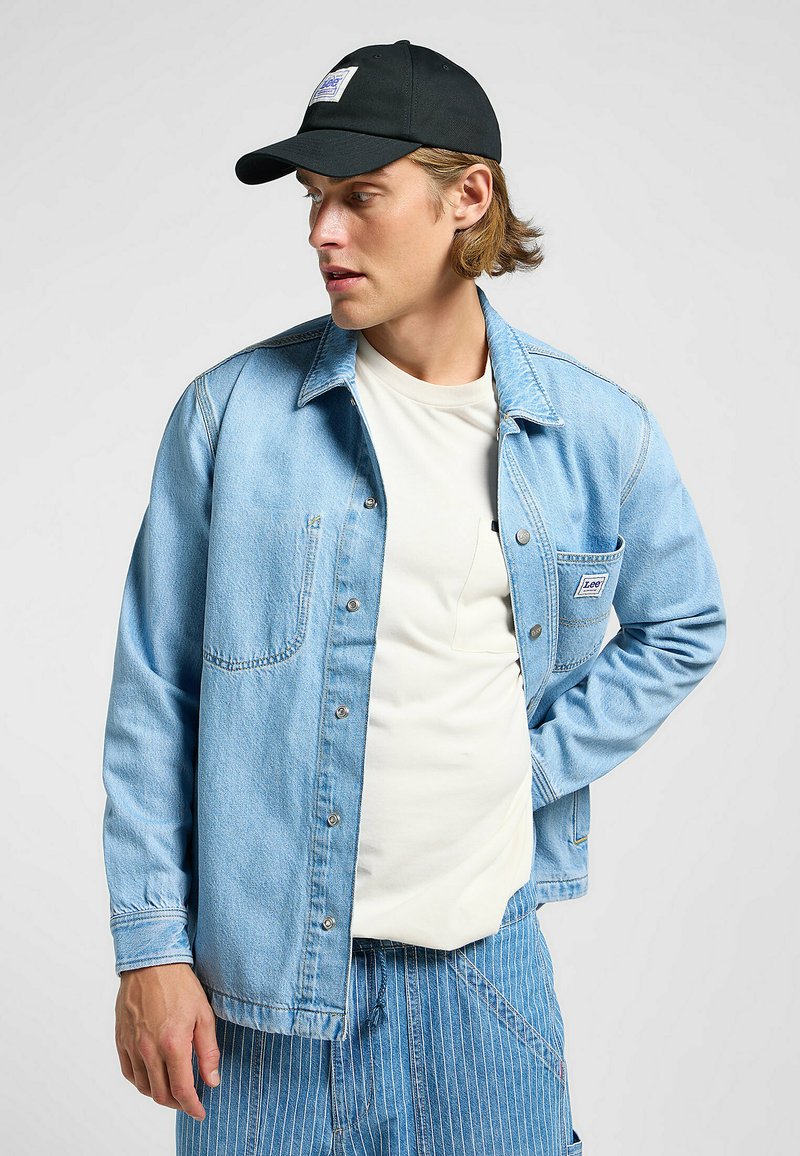 Lys blå denim overskjorte med frontknapper og lomme, parret med en hvid t-shirt og stribede blå denimbukser, toppet med en sort kasket.