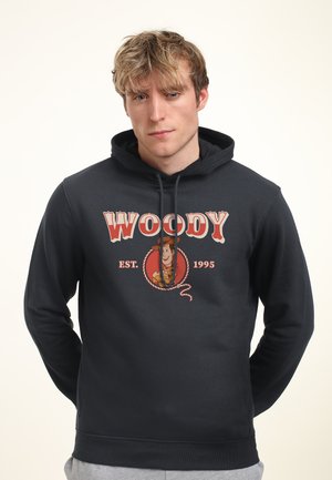 Jonge man draagt een zwarte hoodie met de tekst "Woody Est. 1995" en een cartoonfiguur op de voorkant, staand tegen een effen achtergrond.