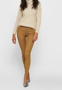 Tan skinny jeans met een gladde textuur, gecombineerd met een beige coltrui. Persoon draagt patroonrijke, puntige hakken.