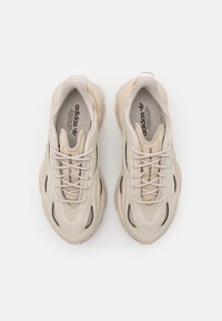 Beige sportskor med en klumpig design, med meshpaneler, tjocka sulor och platta snören på en slät yta.