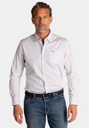 Wit button-up shirt van stof met een subtiel marineblauw geometrisch patroon, enkele borstzak en een rode logo-accent. Ggedragen met blauwe jeans.