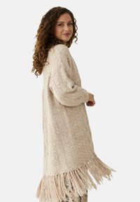 Cárdigan de punto beige con un patrón texturizado, que presenta un dobladillo con flecos y mangas sueltas con canalé. Tiene un ajuste holgado y un diseño de longitud media.