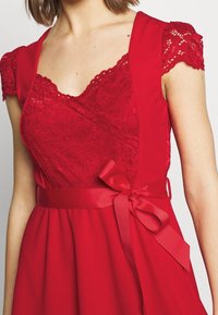 Robe rouge avec des détails en dentelle au corsage, des manches courtes, une taille cintrée avec un nœud en ruban et une jupe évasée.