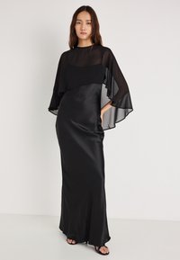 Černé večerní šaty s průhlednou vrstvou, dlouhými rukávy typu netopýr a přiléhavým střihem. Hladká saténová textura, délka až na zem.