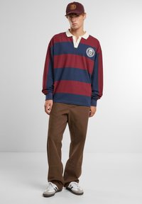 Langärmliges Rugby-Shirt in weinrot und marineblauen Streifen, mit einem weißen Kragen und gesticktem Wappen, kombiniert mit braunen weiten Hosen und Turnschuhen.