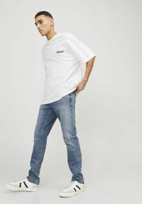 Camiseta de algodón blanca con mangas cortas, que presenta un pequeño logo impreso en negro. Se lleva con jeans desgastados de color azul claro y zapatillas blancas.